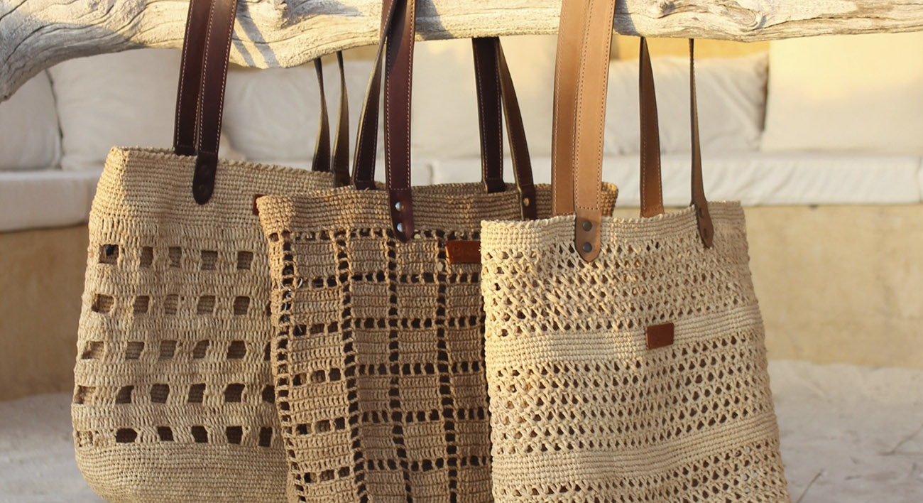 Photo de trois cabas moyens en raphia naturel et avec des motifs différents, points au crochet avec des petits trous, les sacs sont accrochés sur une branche par leur anses solides en raphia tressé ou en cuir véritable.