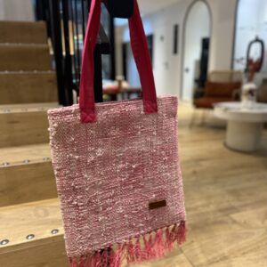 Photo d'un totebag, tissage raphia et coton, tissage traditionnel de Madagascar, couleur fuschia. Sur la base du totebag, il y a des franges en raphia, le totebag est accroché à une rampe d'escalier, il est muni d'une grande anse en tissu couleur fuschia et blanc. Style bohème.