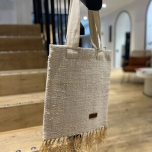 Photo d'un totebag, tissage raphia et coton, tissage traditionnel de Madagascar, couleur blanc. Sur la base du totebag, il y a des franges en raphia, le totebag est accroché à une rampe d'escalier, il est muni d'une grande anse en tissu couleur blanc. Style bohème.