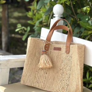 Photo d'un sac moyen cabas Livia en raphia crocheté couleur naturelle, posé sur un banc, artisanat de madagascar, porté main par une femme à la plage, ambiance vacances assurée