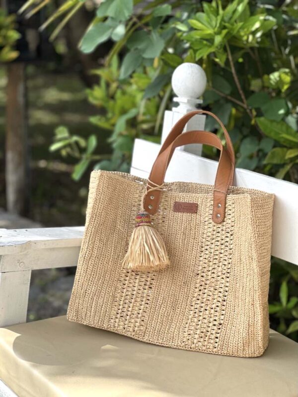 Photo d'un sac moyen cabas Livia en raphia crocheté couleur naturelle, posé sur un banc, artisanat de madagascar, porté main par une femme à la plage, ambiance vacances assurée