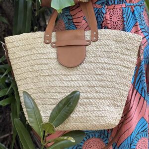 Photo d'une femme debout tenant à on avant-bras un panier en raphia ISALO couleur blanc écru. Le panier est muni d'une petite anse pour porté main, fermé par un rabat en cuir naturel couleur camel. Idéale également pour la ville, panier façon fourre-tout en raphia, artisanat de Madagascar.