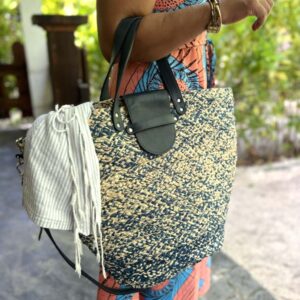 Photo d'une femme debout portant à son avant-bras un panier en raphia ISALO couleur bleu chiné, avec un fouta qui dépasse du panier. Le panier est muni d'une petite anse pour porté main, fermé par un rabat en cuir naturel couleur bleu marine. Idéale également pour la ville, panier façon fourre-tout en raphia, artisanat de Madagascar.