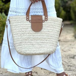 Photo d'une femme debout tenant dans ses mains un panier en raphia ISALO couleur blanc écru. Le panier est muni d'une petite anse pour porté main, fermé par un rabat en cuir naturel couleur camel. Idéale également pour la ville, panier façon fourre-tout en raphia, artisanat de Madagascar.