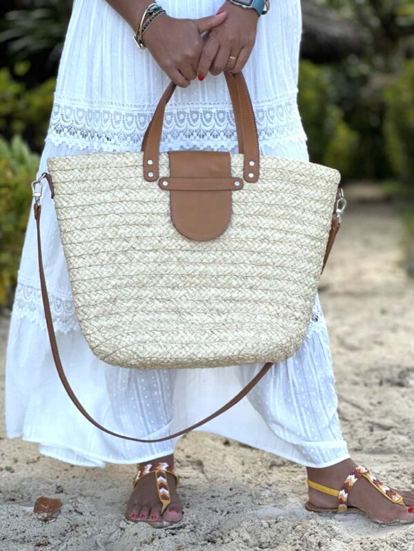 Photo d'une femme debout tenant dans ses mains un panier en raphia ISALO couleur blanc écru. Le panier est muni d'une petite anse pour porté main, fermé par un rabat en cuir naturel couleur camel. Idéale également pour la ville, panier façon fourre-tout en raphia, artisanat de Madagascar.