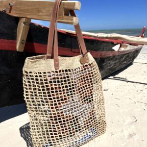 Photo d'un grand sac de plage LALAO en raphia naturel point filet, accroché à une pirogue immobilisé sur une plage de sable, avec une anse en cuir véritable couleur camel. Ambiance vacances d'été, plage, océan.