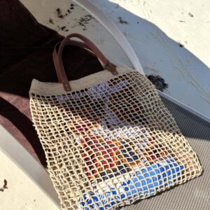 Photo d'un grand sac de plage LALAO en raphia naturel point filet, posé sur un transat à la plage, avec une anse en cuir véritable couleur camel. Ambiance vacances d'été, plage, océan.