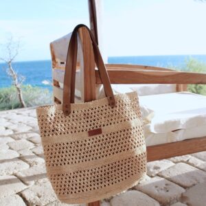 Photo d'un sac en raphia naturel accroché à un fauteuil sur une terrasse extérieure, sac MEVA avec point au crochet motif petits trous, ambiance vacances en bord de mer.