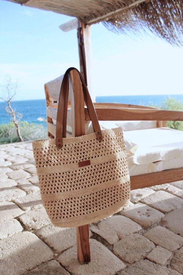 Photo d'un sac en raphia naturel accroché à un fauteuil sur une terrasse extérieure, sac MEVA avec point au crochet motif petits trous, ambiance vacances en bord de mer.