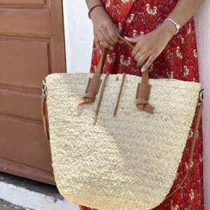 Photo d'une jeune femme appuyée contre un mur avec un grand panier en raphia tressé porté main panier raphia avec une grande bandoulière en cuir naturel couleur camel.