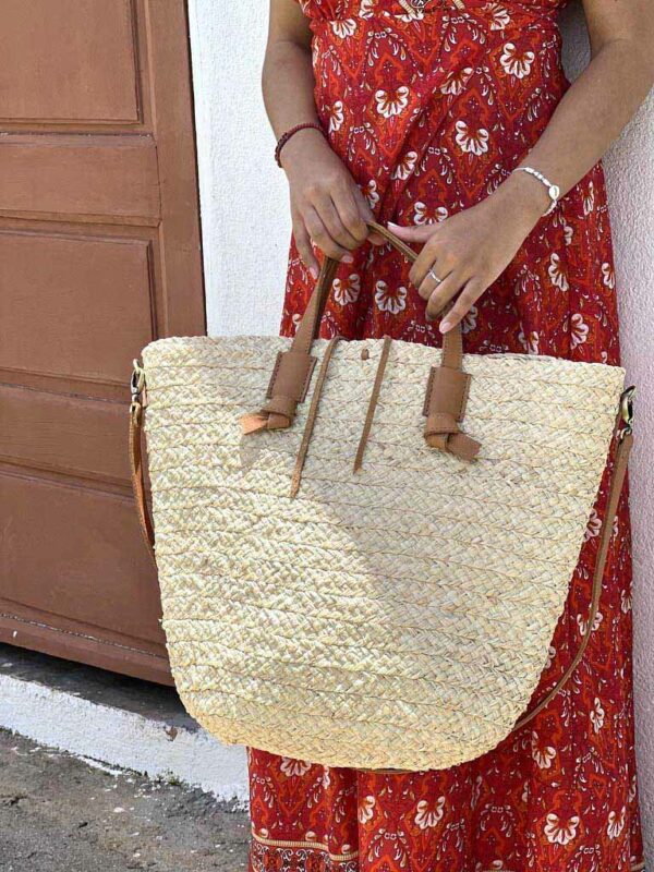 Photo d'une jeune femme appuyée contre un mur avec un grand panier en raphia tressé porté main panier raphia avec une grande bandoulière en cuir naturel couleur camel.