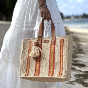 Photo d'une femme à la plage avec le cabas MAYA de taille moyenne en raphia, porté main, couleur naturel avec des bandes rouges et jaunes, artisanat de MADAGASCAR, cabas fait main au crochet, idéal pour l'été.