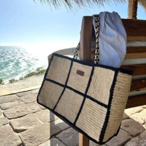 Photo d'un grand cabas de plage MARINA accroché à un accoudoir de fauteuil face à la mer, ambiance vacances d'été, sac en raphia couleur naturel, motif grand carré damier avec bordure noir, anse en raphia tressé couleur noir et naturel