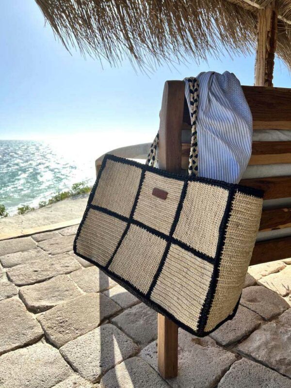 Photo d'un grand cabas de plage MARINA accroché à un accoudoir de fauteuil face à la mer, ambiance vacances d'été, sac en raphia couleur naturel, motif grand carré damier avec bordure noir, anse en raphia tressé couleur noir et naturel