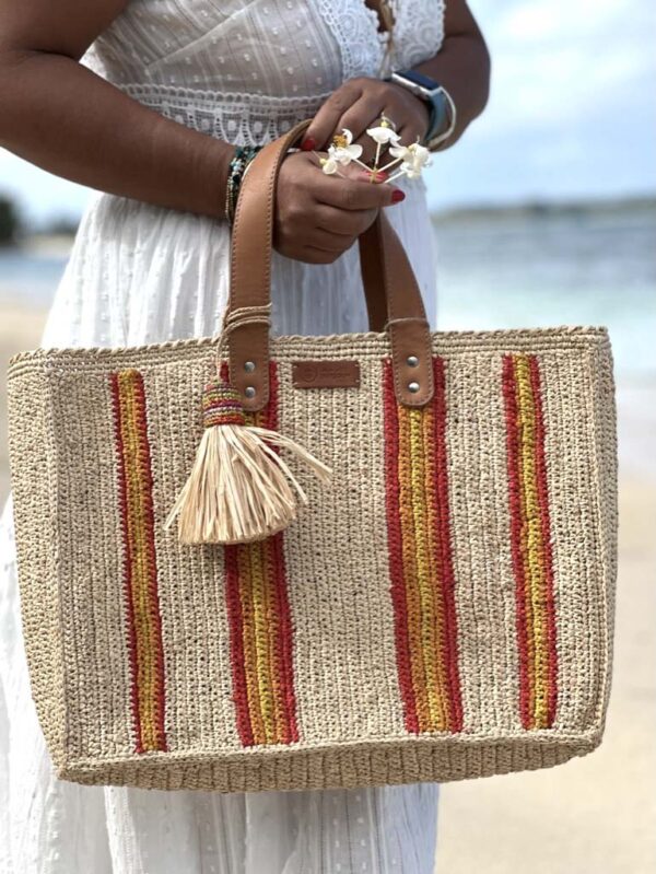 Photo du cabas moyen Maya de Madagascar , porté main par une femme à la plage, artisanat de Madagascar, motif rayures rouges et jaunes.