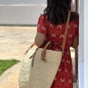 Photo d'une jeune femme appuyée contre un mur avec un grand panier en raphia tressé porté épaule, panier raphia avec une grande bandoulière en cuir naturel couleur camel.