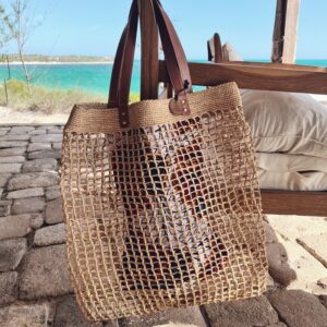 Photo d'un grand sac de plage LALAO en raphia naturel, point filet, accroché à un fauteuil avec une anse en cuir véritable couleur camel, Photo d'une fille qui tient à sa main un totebag en raphia couleur thé, un totebag super léger et original, en raphia motif macramé, apporte un style bohème à une tenue estivale. Ambiance vacances d'été, plage, océan.