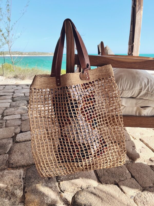 Photo d'un grand sac de plage LALAO en raphia naturel, point filet, accroché à un fauteuil avec une anse en cuir véritable couleur camel, Photo d'une fille qui tient à sa main un totebag en raphia couleur thé, un totebag super léger et original, en raphia motif macramé, apporte un style bohème à une tenue estivale. Ambiance vacances d'été, plage, océan.