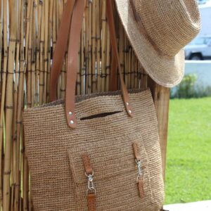 Photo d'un sac de ville en raphia crocheté couleur thé, accroché à une cloture en bambou à côté d'un chapeau en raphia, le sac comporte une poche extérieure fermée par deux mousquetons. Un sac en raphia bien pratique pour la ville. Artisanat de Madagascar.