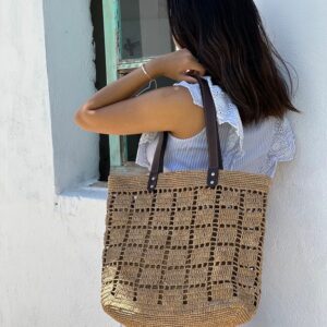 Photo d'une femme appuyé contre un mur portant un cabas NIRINA en raphia de taille moyenne sur une épaule, grande ouverture, muni d'une pochette entièrement en raphia à l'intérieur, la pochette est fermée par une fermeture, doublée en tissu coton, elle est tenue par un mousqueton et est amovible, peut être enlevée du cabas. Cabas fait main à Madagascar. couleur thé, motif aéré par des petits carrés sur tout l'ensemble du cabas.