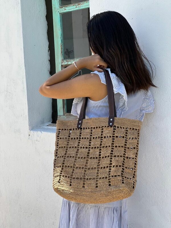 Photo d'une femme appuyé contre un mur portant un cabas NIRINA en raphia de taille moyenne sur une épaule, grande ouverture, muni d'une pochette entièrement en raphia à l'intérieur, la pochette est fermée par une fermeture, doublée en tissu coton, elle est tenue par un mousqueton et est amovible, peut être enlevée du cabas. Cabas fait main à Madagascar. couleur thé, motif aéré par des petits carrés sur tout l'ensemble du cabas.