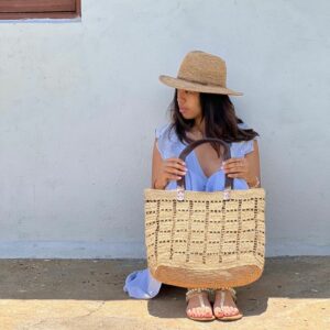 Photo d'une femme assise par terre, tenant dans ses mains un cabas NIRINA en raphia de taille moyenne, grande ouverture, muni d'une pochette entièrement en raphia à l'intérieur, la pochette est fermée par une fermeture, doublée en tissu coton, elle est tenue par un mousqueton et est amovible, peut être enlevée du cabas. Cabas fait main à Madagascar. couleur thé, motif aéré par des petits carrés sur tout l'ensemble du cabas.