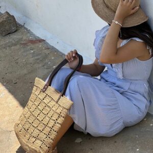 Photo d'une femme assise par terre, tenant dans sa main un cabas NIRINA en raphia de taille moyenne, grande ouverture, muni d'une pochette entièrement en raphia à l'intérieur, la pochette est fermée par une fermeture, doublée en tissu coton, elle est tenue par un mousqueton et est amovible, peut être enlevée du cabas. Cabas fait main à Madagascar. couleur thé, motif aéré par des petits carrés sur tout l'ensemble du cabas.