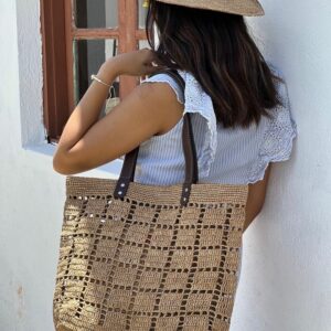 Photo d'une femme appuyé contre un mur portant un cabas NIRINA en raphia de taille moyenne sur une épaule, grande ouverture, muni d'une pochette entièrement en raphia à l'intérieur, la pochette est fermée par une fermeture, doublée en tissu coton, elle est tenue par un mousqueton et est amovible, peut être enlevée du cabas. Cabas fait main à Madagascar. couleur thé, motif aéré par des petits carrés sur tout l'ensemble du cabas.